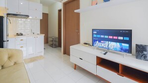 A modern living room with a flat-screen TV displaying a movie, a sofa, a coffee table, and a kitchen area with white cabinets and a stove. - Cozy and Simply Look 2BR Podomoro Golf View Apartment (Gunung Putri)