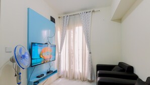 Ein Wohnzimmer mit an einer blauen Wand montiertem Fernseher, einem Standventilator, einem schwarzen Sofa und einem Fenster mit durchscheinenden Vorhängen.