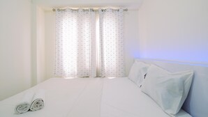 Ein ordentlich bezogenes Bett mit weißen Leinen und Kissen, ein Fenster mit geometrisch gemusterten Vorhängen und ein Kopfteil mit einem Lichtschalter.