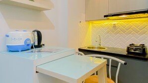 Una cocina compacta con encimera blanca, un fregadero y un hervidor. Hay una pequeña mesa con una silla y una planta decorativa sobre una repisa.