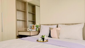 Una cama bien tendida con una manta morada, una mesita de noche con un florero y un espejo que refleja la habitación.