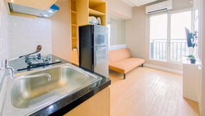 A modern kitchen with a sink, a black refrigerator, and a built-in oven. There is a beige sofa and a window with a view of the outside. - Good Place 2BR at Podomoro Golf View Apartment (Gunung Putri)