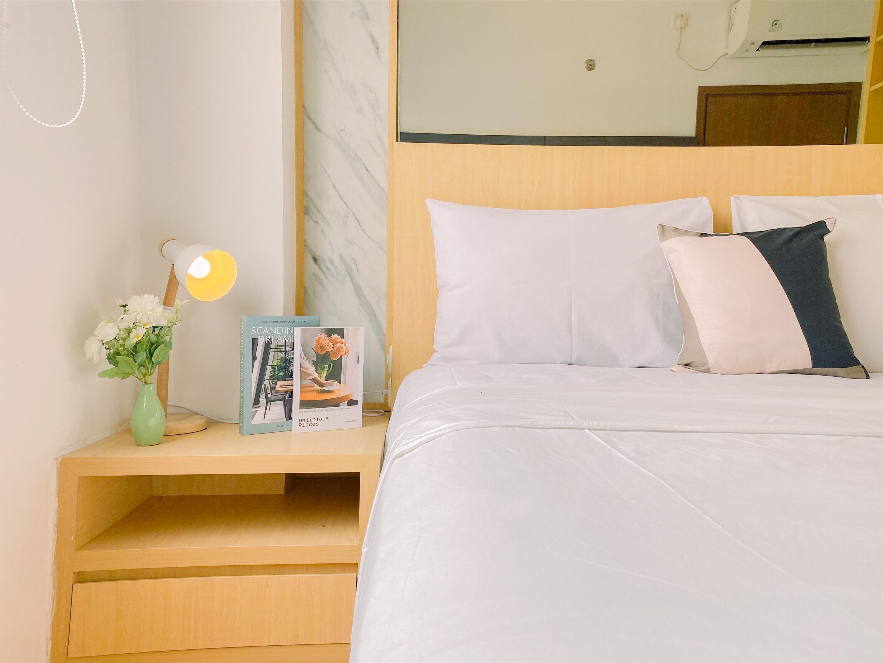 A neatly made bed with white bedding, a bedside table with a lamp, a vase with flowers, and two books.