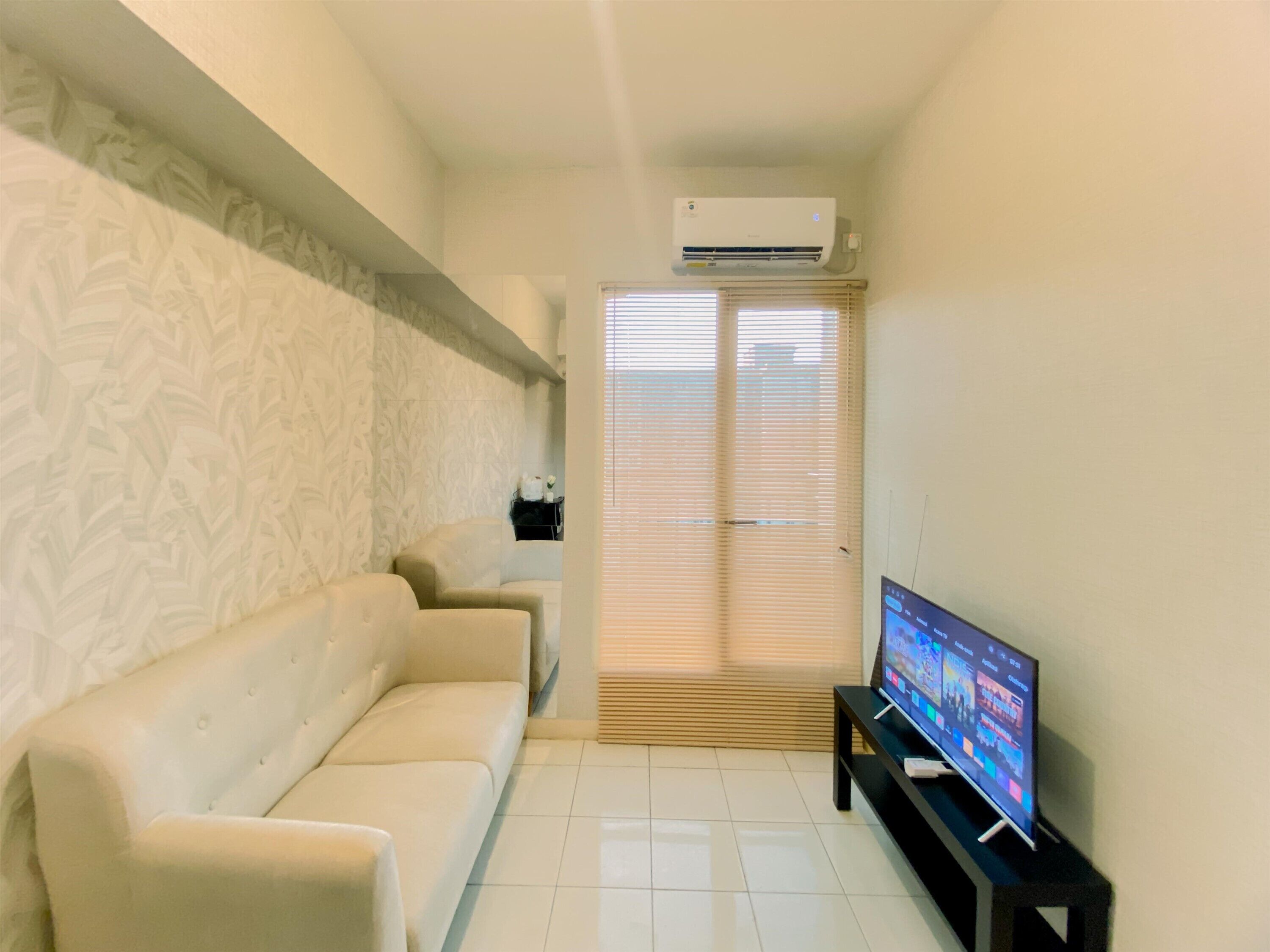 A living room with a white sofa, a flat-screen TV on a stand, and a window with blinds.