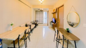 A dining area with a long wooden table and chairs, a sofa, a TV, and a mirror on the wall. - Well Furnished and Comfy 3BR Podomoro Golf View Apartment (Gunung Putri)