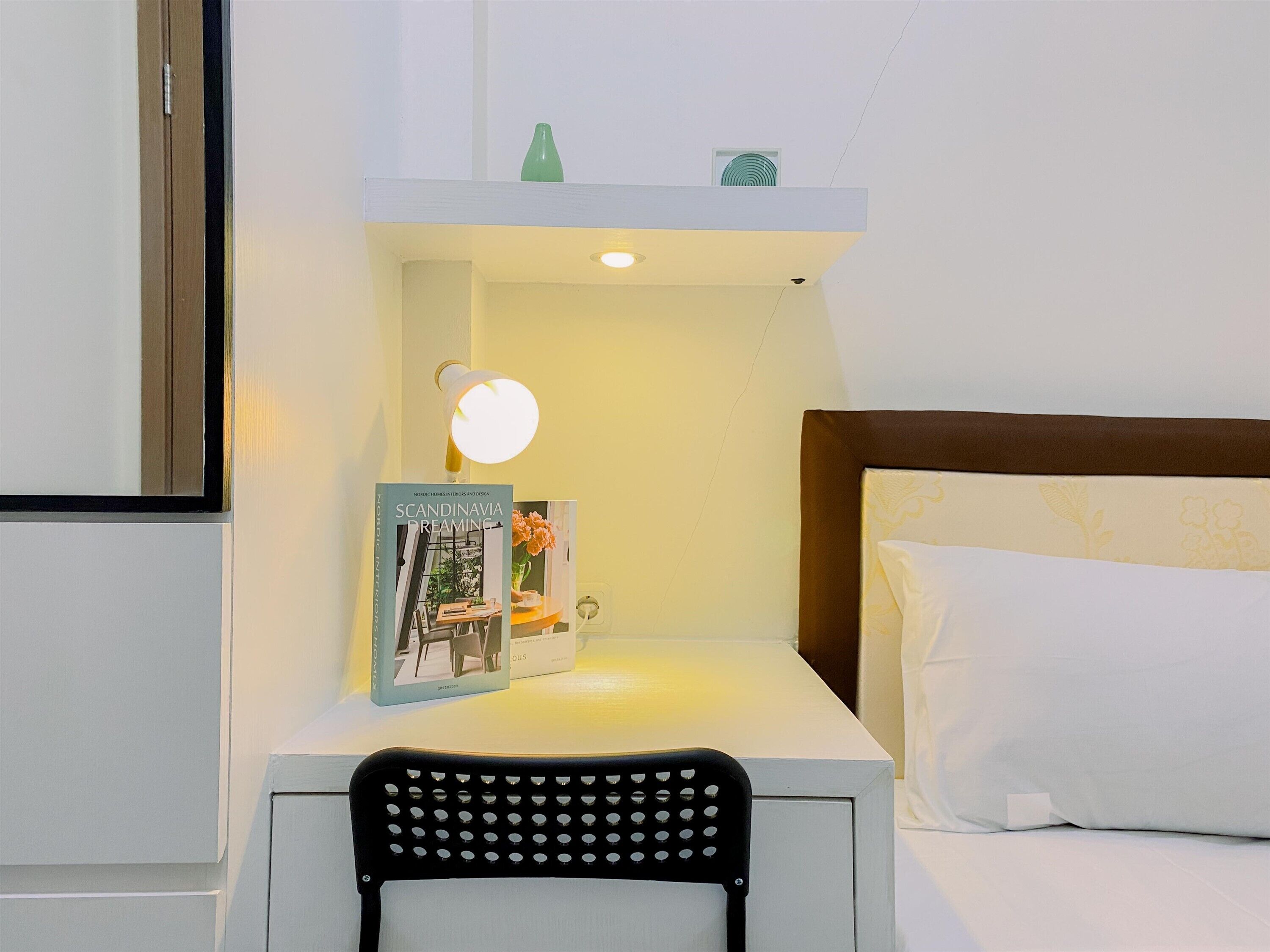 A neatly arranged hotel room with a bedside table, a chair, a bed with white bedding, a book titled "Scandinavian Dreaming," and a small green bottle on the nightstand.