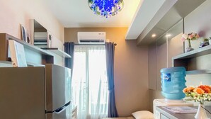 Una habitación de hotel compacta con kitchenette, una cama con colchón color crema, un dispensador de agua y una pequeña mesa con un florero.