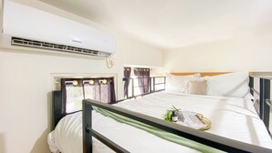 A hotel room with a bed, a window with curtains, an air conditioning unit, and a bedside table with a bottle and a tray. - Tranquil and Homey Studio at Podomoro Golf View Apartment (Gunung Putri)