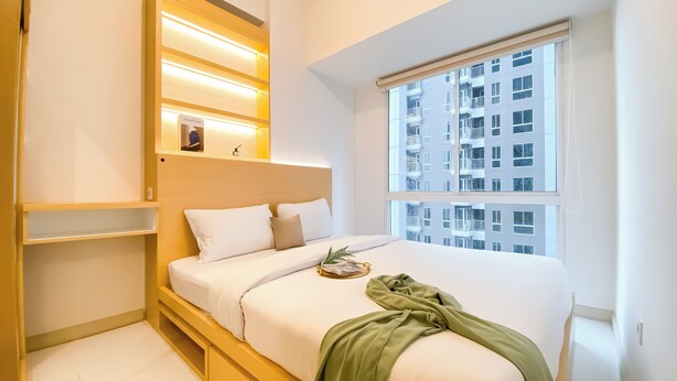 Une chambre moderne avec un grand lit, des étagères intégrées, une fenêtre donnant sur un immeuble et une petite plante posée sur le lit.