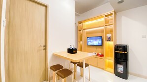 Une chambre d’hôtel moderne avec une porte en bois, une petite table avec deux chaises, un téléviseur fixé au mur, un distributeur d’eau et une étagère avec un livre.