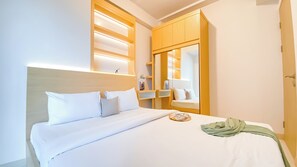 A neatly made bed with white linens, a bedside table with a book and a glass, and a mirror reflecting part of the room. - Comfy and Modern Look 2BR Apartment at Tokyo Riverside PIK 2 (Teluknaga)