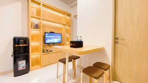 A modern hotel room with a wooden desk, two chairs, a flat-screen TV, a minibar, and a minibar fridge. - Comfy and Modern Look 2BR Apartment at Tokyo Riverside PIK 2 (Teluknaga)