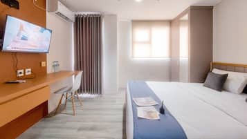 Quarto de hotel com cama, escrivaninha com cadeira, televisão e janela com cortinas.