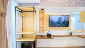 A hotel room with a TV mounted on the wall displaying a scenic mountain view, a wooden shelf unit with hangers, a small table with a remote control, and a minibar. - Minimalist and Homey Studio at Tokyo Riverside PIK 2 Apartment (Teluknaga)