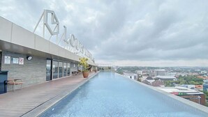 A rooftop infinity pool with a view of the city, a modern building with 'LOUIS' signage, and a clear sky. - Good Homey Studio Apartment at Warhol (W/R) Residences (Semarang)