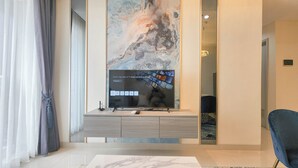 A modern living room with a large flat-screen TV on a wooden console, a marble-patterned wall, and a blue chair. - Wonderful 2BR with City View at Grand Sungkono Lagoon Apartment (Surabaya)