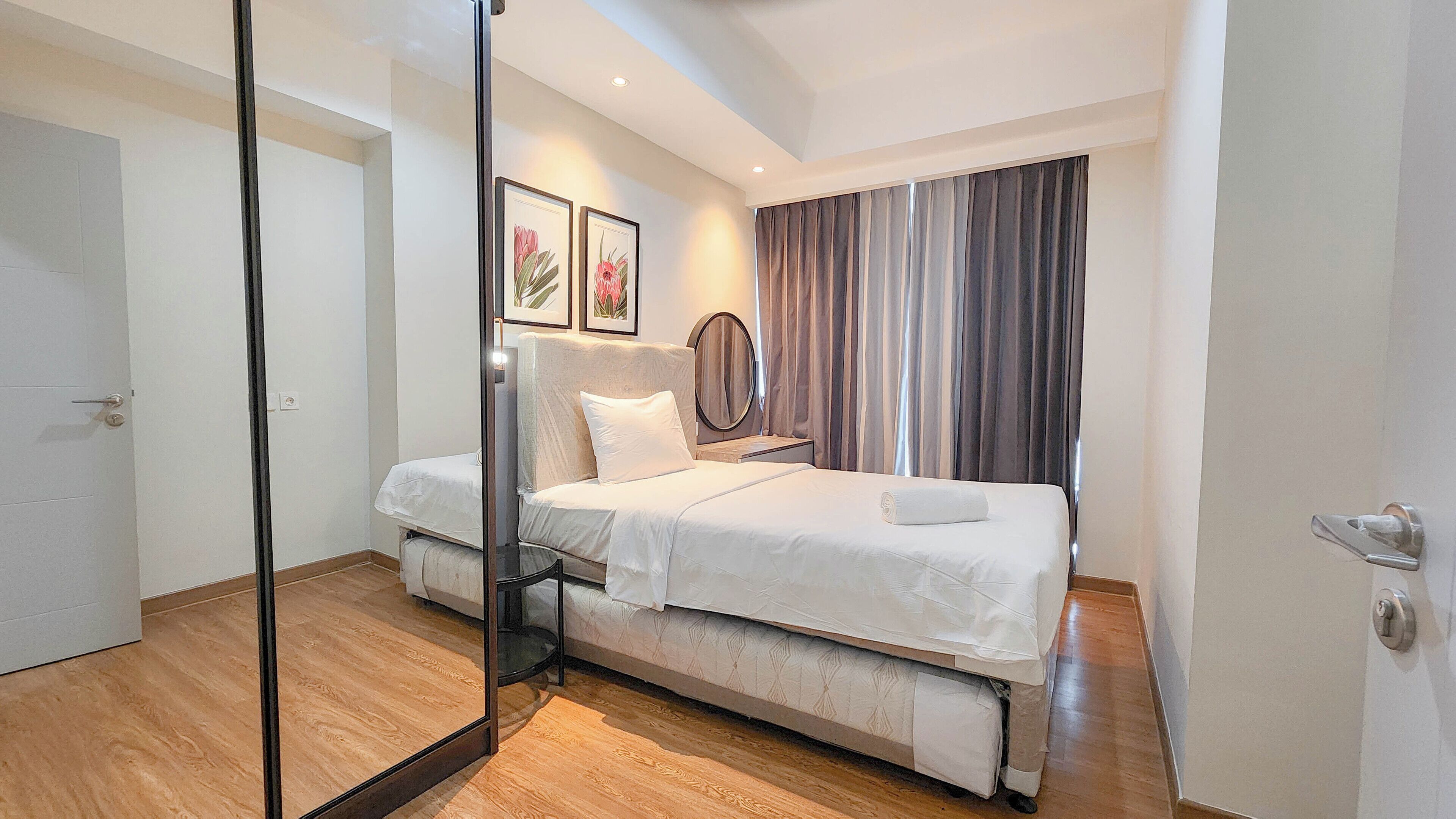 A modern bedroom with a bed, bedside table, and a mirror. There is a door and a window with curtains.