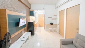 Una sala moderna con un sofá, un televisor montado en la pared, una mesita con un ventilador y una zona de cocina al fondo.