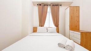 Una cama bien hecha con sábanas blancas y una toalla enrollada, una mesita de noche de madera y un armario de madera en una habitación luminosa y con pocos muebles.
