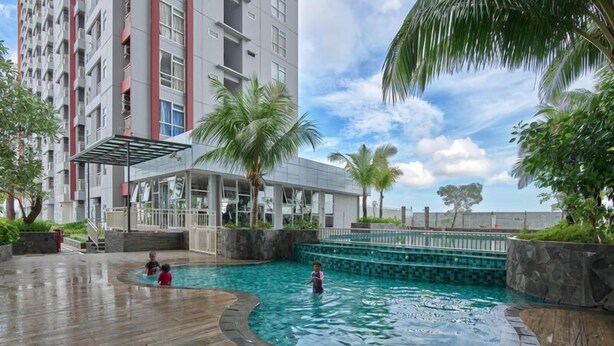 A swimming pool area with a wooden deck, palm trees, and a building in the background. - The Urban 2BR at Vida View Makassar Apartment (Makassar)
