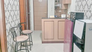 A small kitchen with a wooden cabinet, a sink, and a countertop. There are two stools and a refrigerator. - The Urban 2BR at Vida View Makassar Apartment (Makassar)