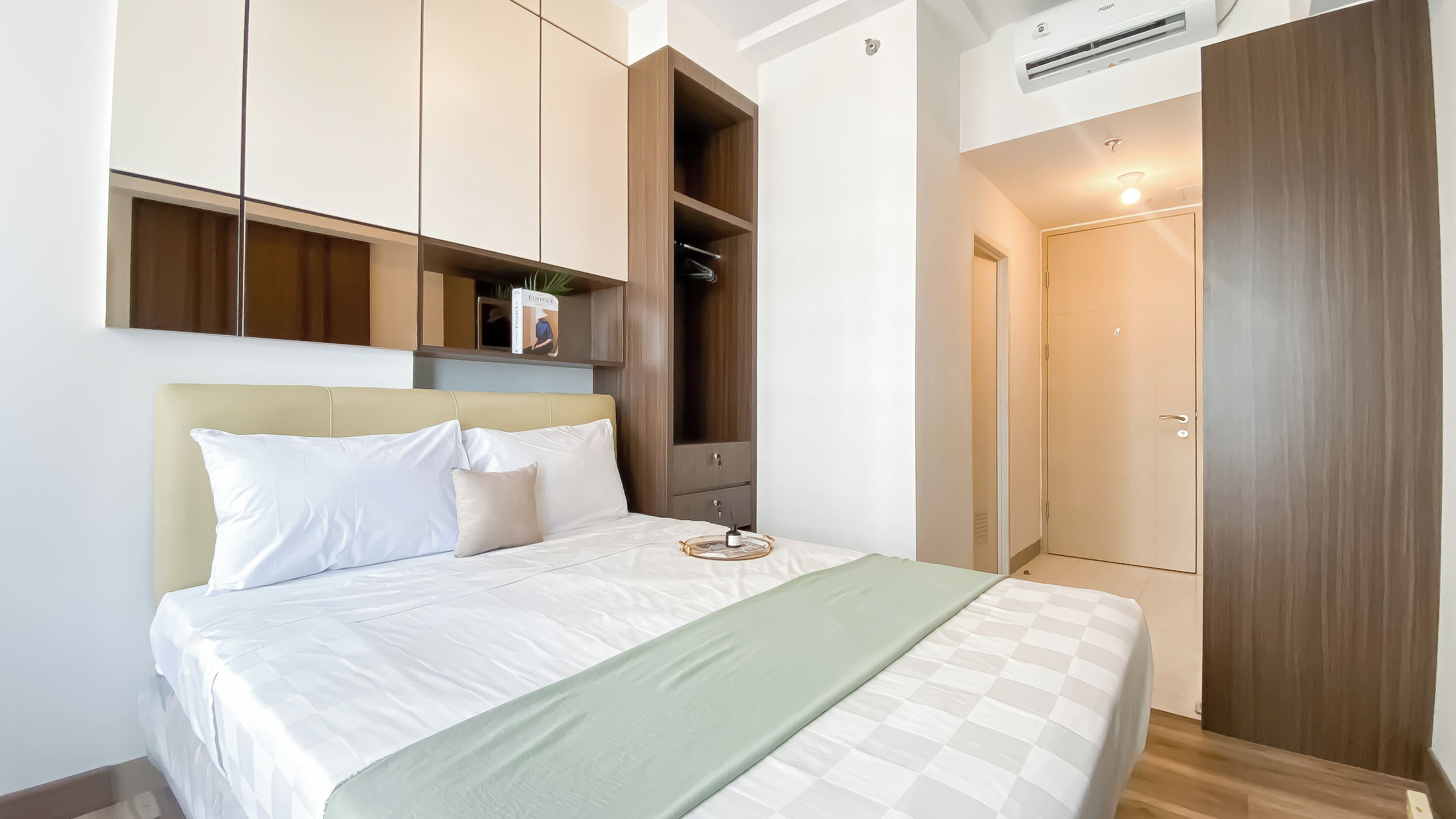 A neatly made bed with white bedding and a green throw, a wooden wardrobe with a mirror, and a small bedside table with a lamp.