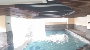An indoor swimming pool with clear water, tiled floor, and a modern architectural design with a sloped ceiling and stone walls. - The Bohemian 2BR at Tamansari La Grande Apartment (Bandung)