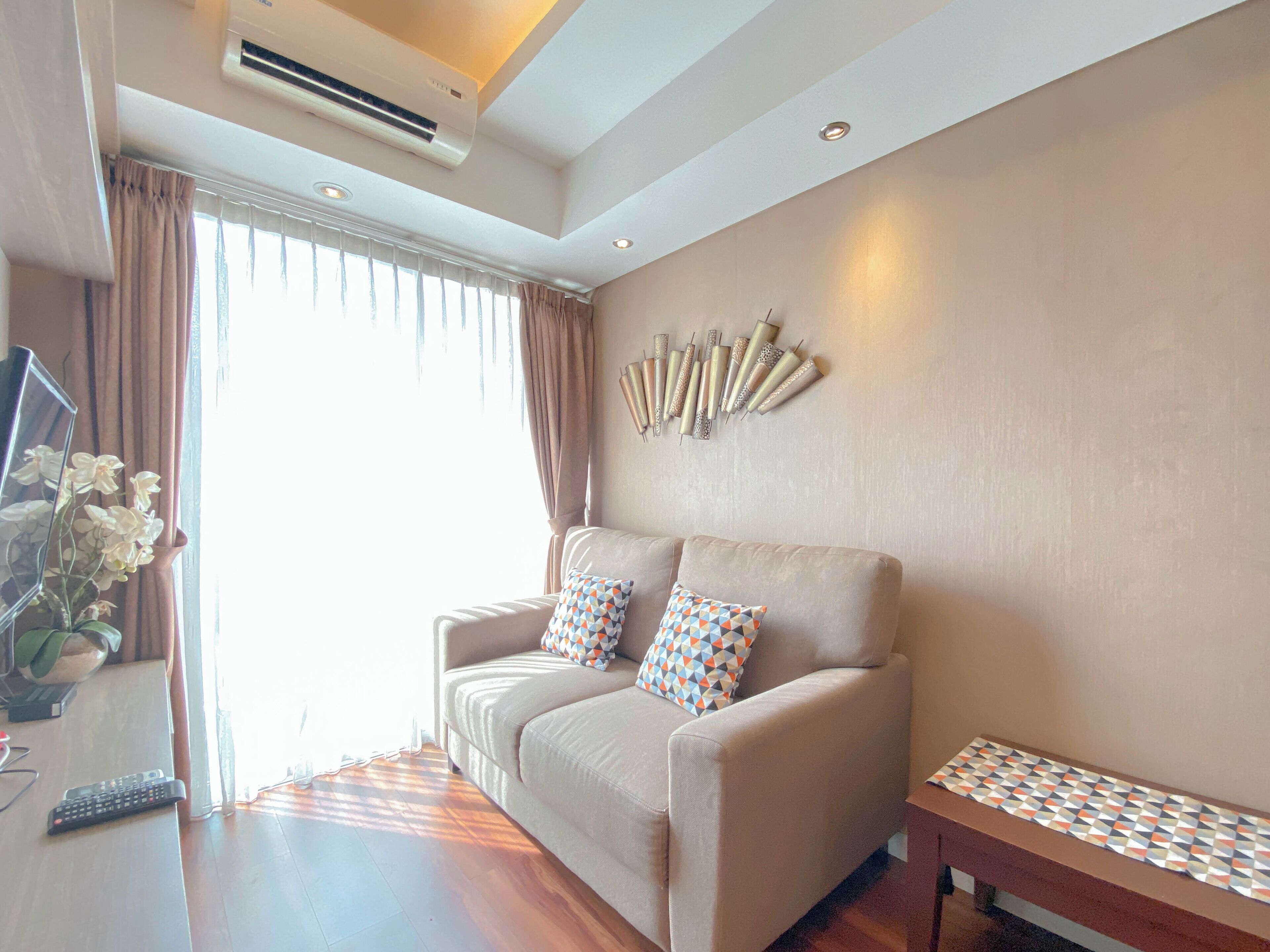 A modern living room with a beige sofa, patterned pillows, a wooden coffee table, and a wall-mounted decorative piece.
