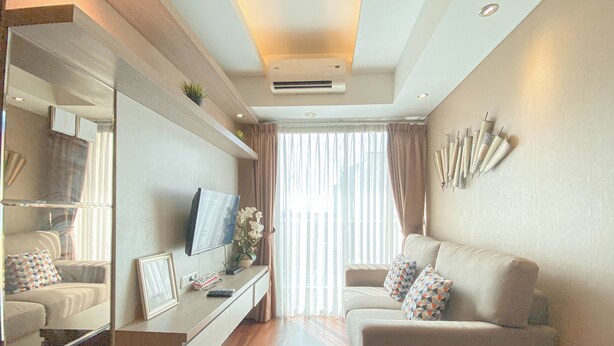 A modern living room with a sofa, a TV, a coffee table, and a large window with curtains. - The Bohemian 2BR at Tamansari La Grande Apartment (Bandung)