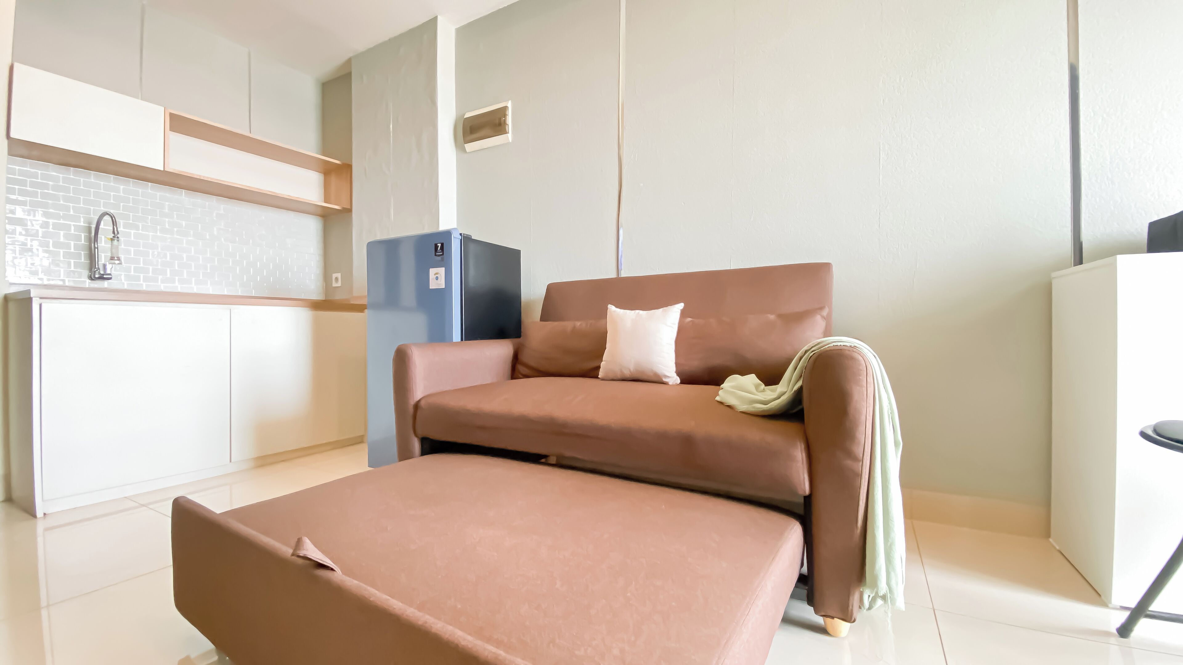 A modern living room with a sofa and ottoman, a kitchen area with a fridge, and a small cabinet.
