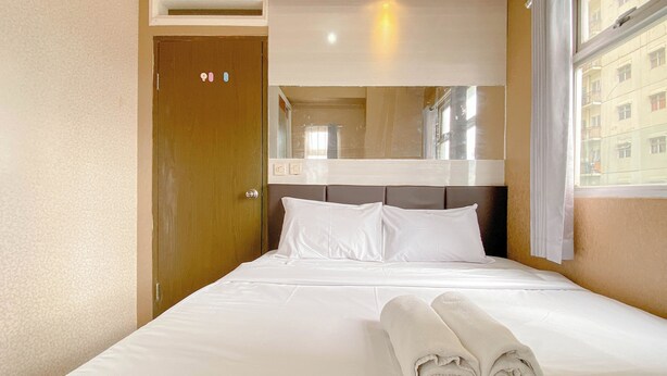 A hotel room with a bed, a wooden door, a mirror above the bed, and a window with a view of buildings. - The Urban Style 2BR at Suites @Metro Apartment (Bandung)