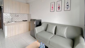 A modern living space with a grey sofa, a kitchen area with wooden cabinets, and a refrigerator. There are three framed pictures on the wall.