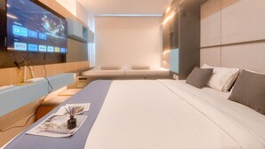 Una habitación de hotel moderna con un televisor de pantalla plana grande montado en la pared, una cama con cubrecama azul y una mesita de noche con una revista y un pequeño jarrón.