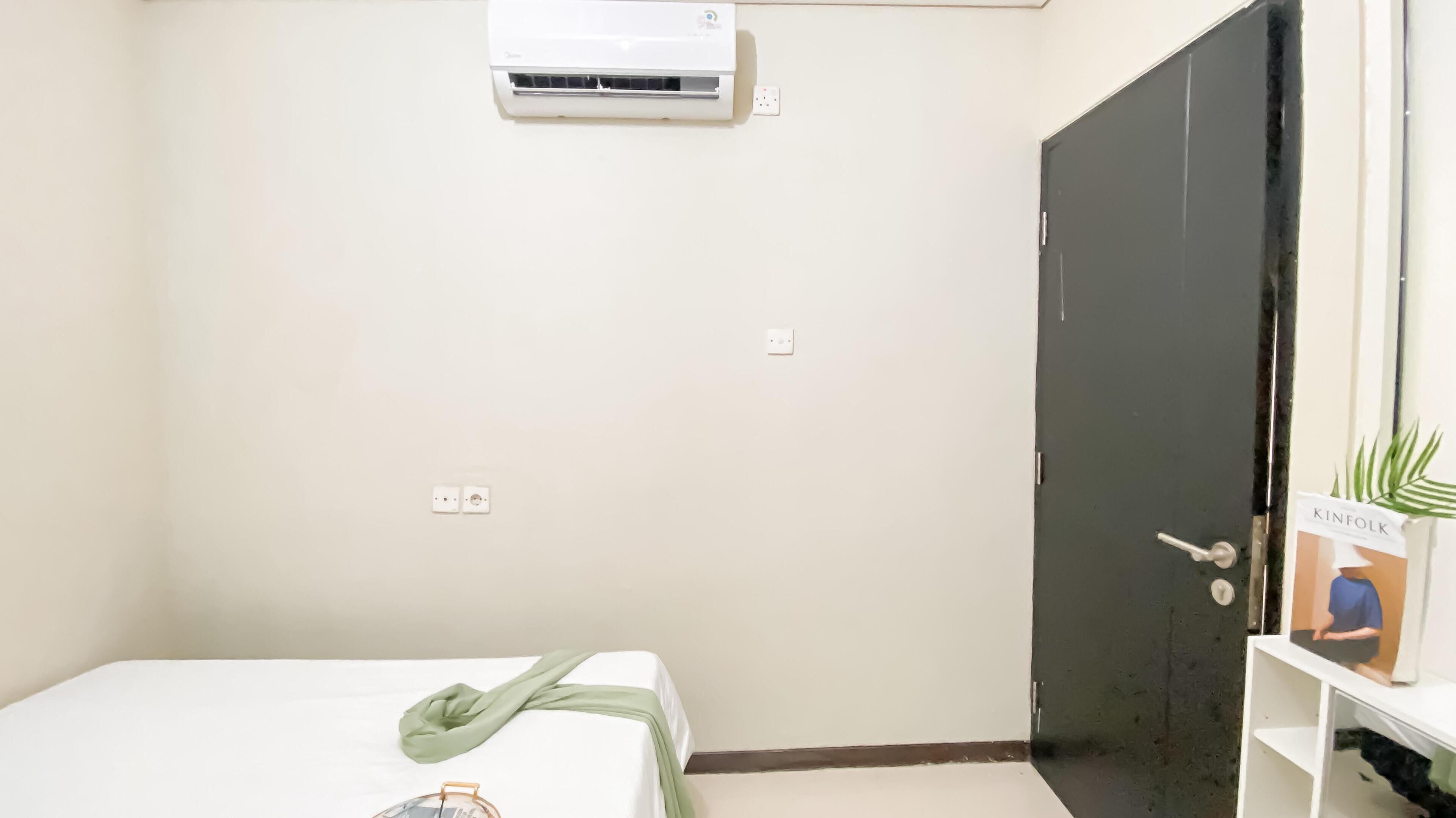 A room with a bed, an air conditioner, a door, and a small shelf with a plant and a poster.