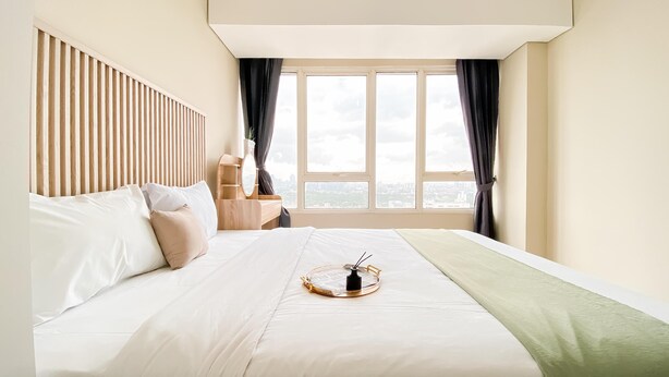 A hotel room with a bed, a bedside table with a mirror, a window with curtains, and a view of the city. - New Furnished and Homey 2BR Apartment Nifarro Park (Jakarta)