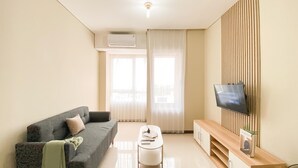 A modern living room with a sofa, a coffee table, a TV mounted on a wooden panel, and a large window with sheer curtains. - New Furnished and Homey 2BR Apartment Nifarro Park (Jakarta)