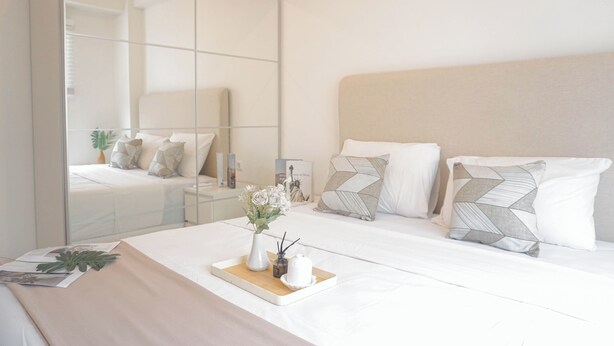 A modern bedroom with a large mirror, a bed with white bedding, and a bedside table with a vase of flowers.
