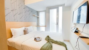 A modern bedroom with a large bed, a wooden headboard, a wall-mounted TV, and a window with blinds. - Cozy Style Studio Apartment at Tokyo Riverside PIK 2 (Teluknaga)