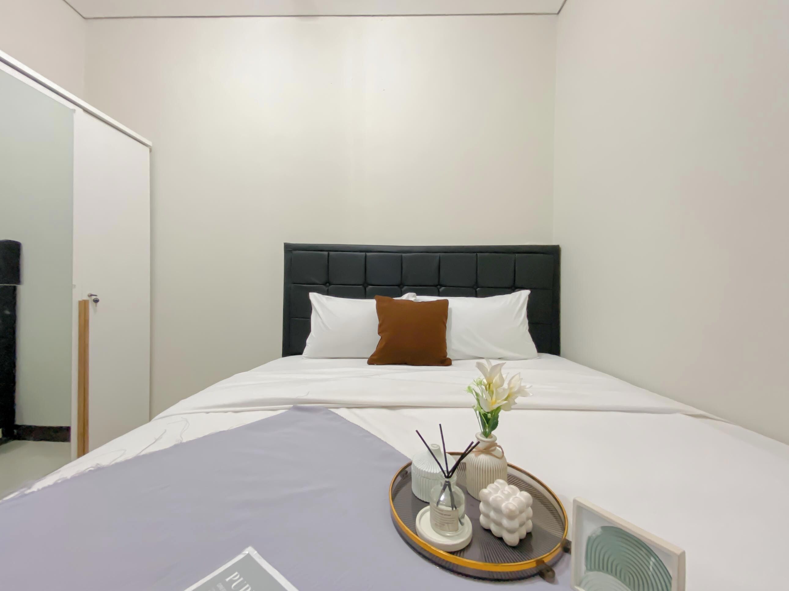 A neatly made bed with white and brown pillows, a dark headboard, and a tray with a vase of flowers and a small decorative item.