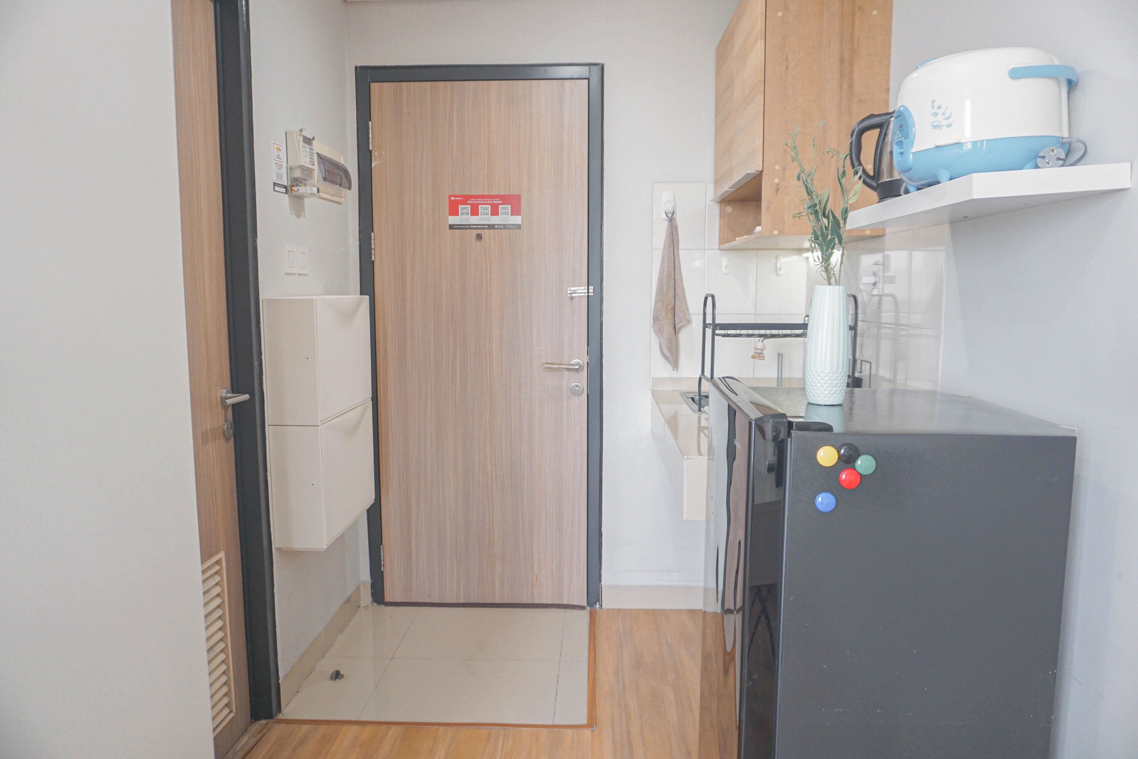 A compact kitchen with a wooden door, a black refrigerator, and a shelf with a kettle and a plant.