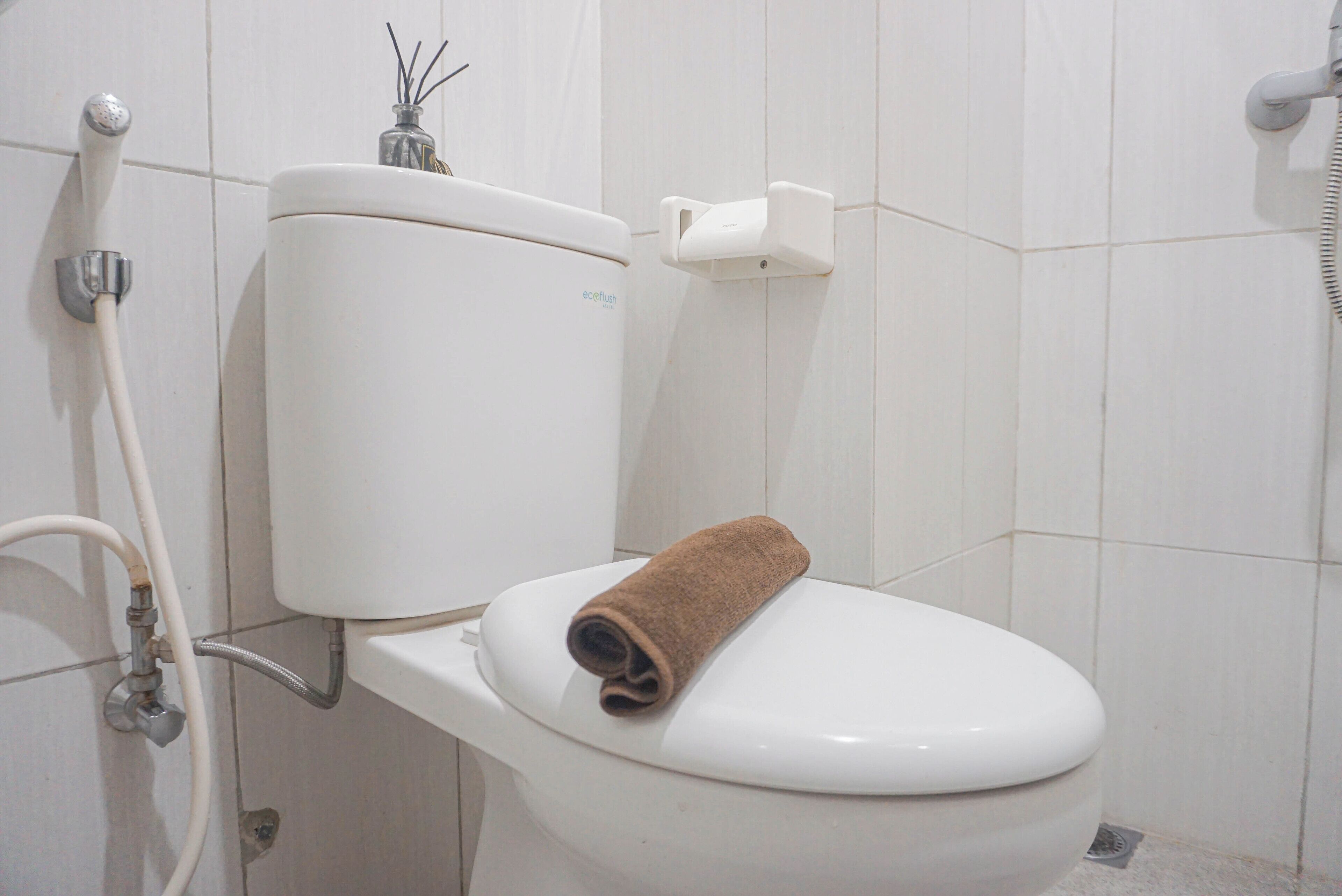 Inodoro blanco con una toalla marrón sobre el asiento, dispensador de jabón para manos y manguera de ducha.