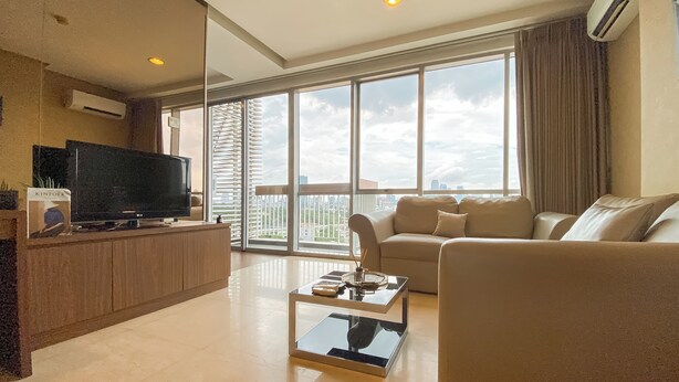 A modern living room with a flat-screen TV, a beige sofa, a glass coffee table, and large windows offering a city view. - Elegant and Spacious Studio at Kemang Mansion Apartment (Jakarta)