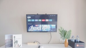 Un téléviseur à écran plat fixé au mur, diffusant le film des Avengers des studios Marvel, une télécommande, un plateau avec une bouteille, un vase avec une plante et deux livres intitulés « Hyde Avenue » et « La Statue de la Liberté ».