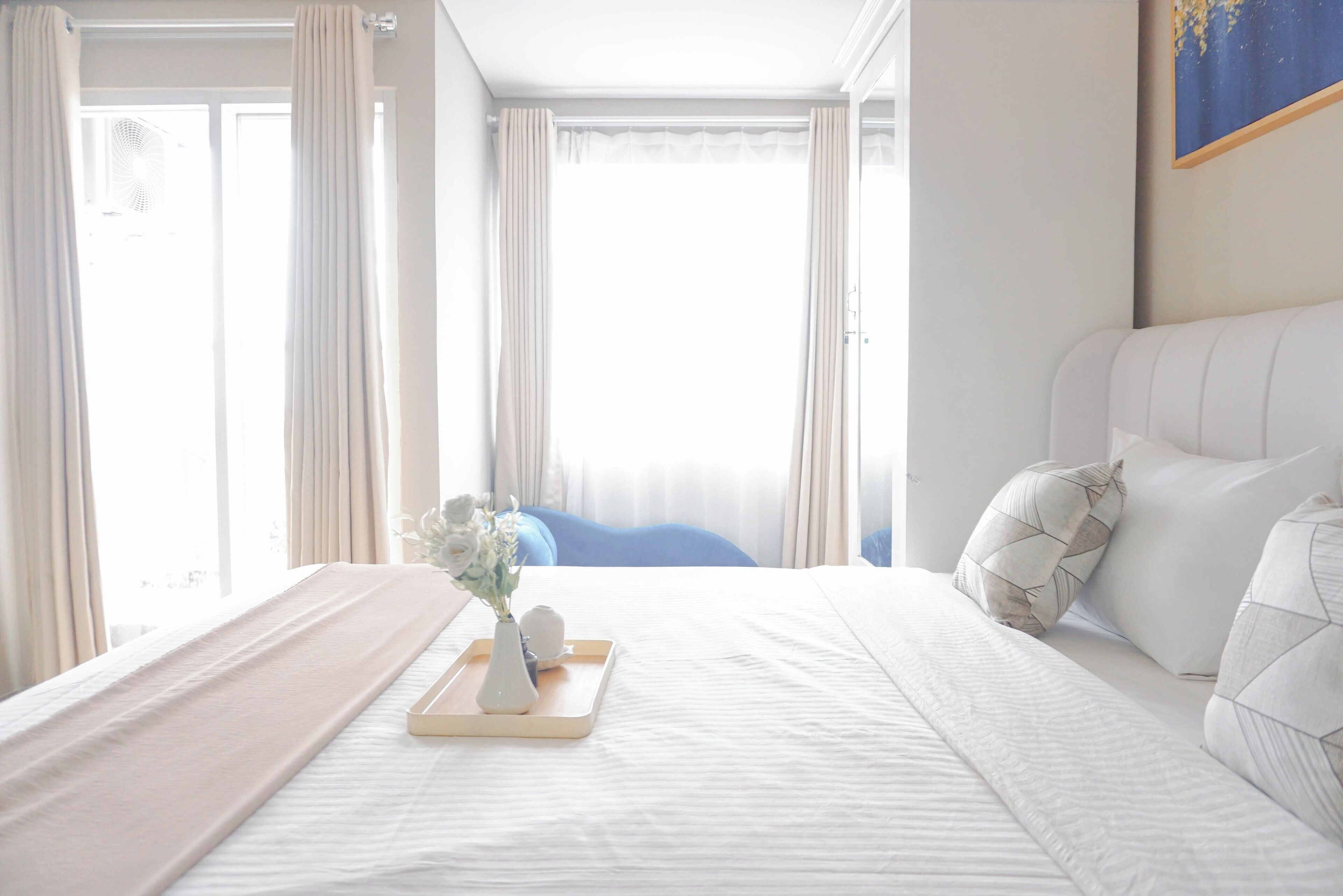 A hotel room with a large bed, a bedside table with a vase of flowers, and a view of the outdoors through sheer curtains.