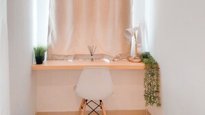 Een kamer met een houten bureau, een witte stoel, een raam met dunne gordijnen, een bureaulamp en een kleine plant in een pot.