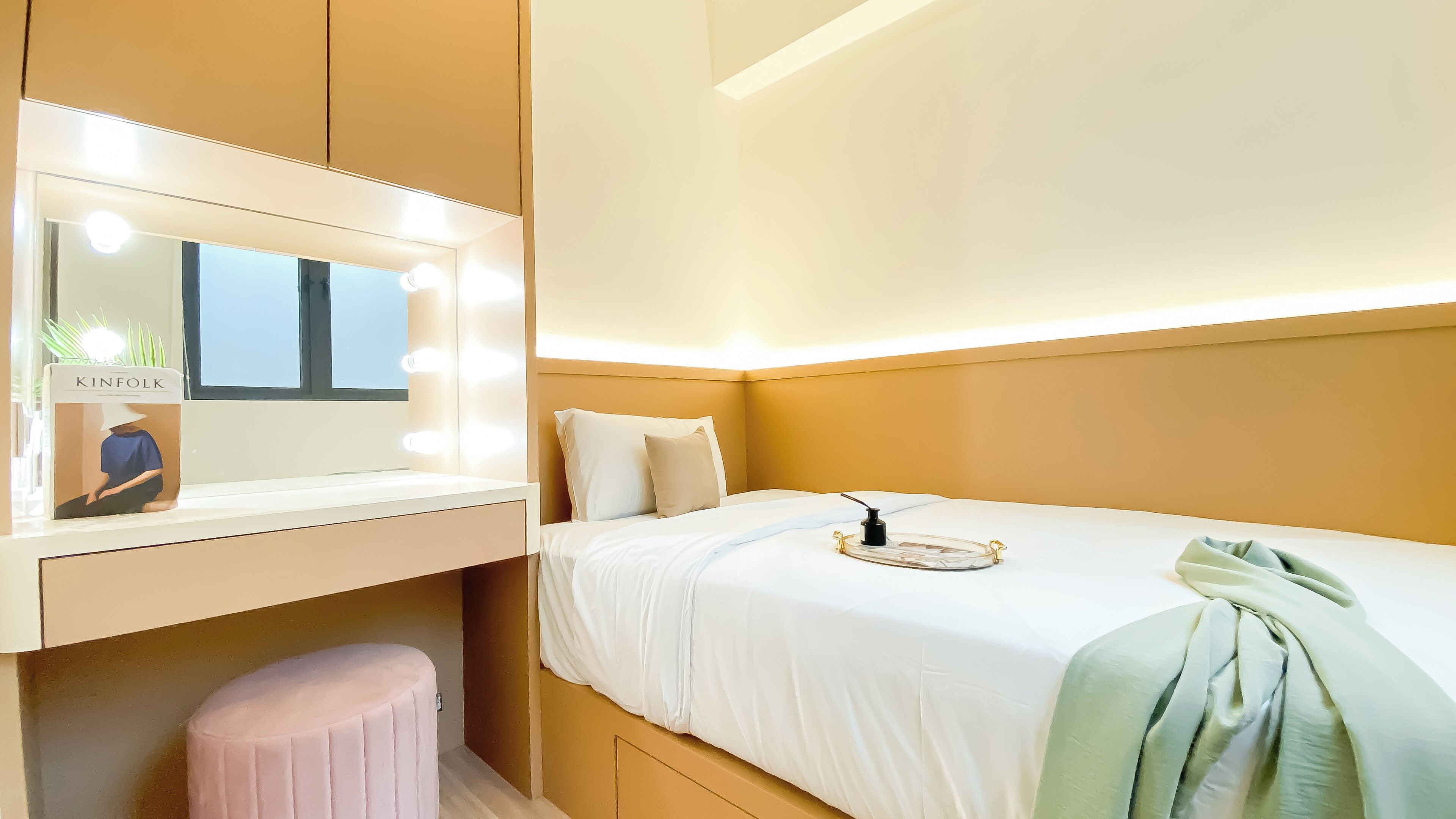 A hotel room with a vanity mirror, a small stool, a bed with white bedding, and a bedside table with a lamp.