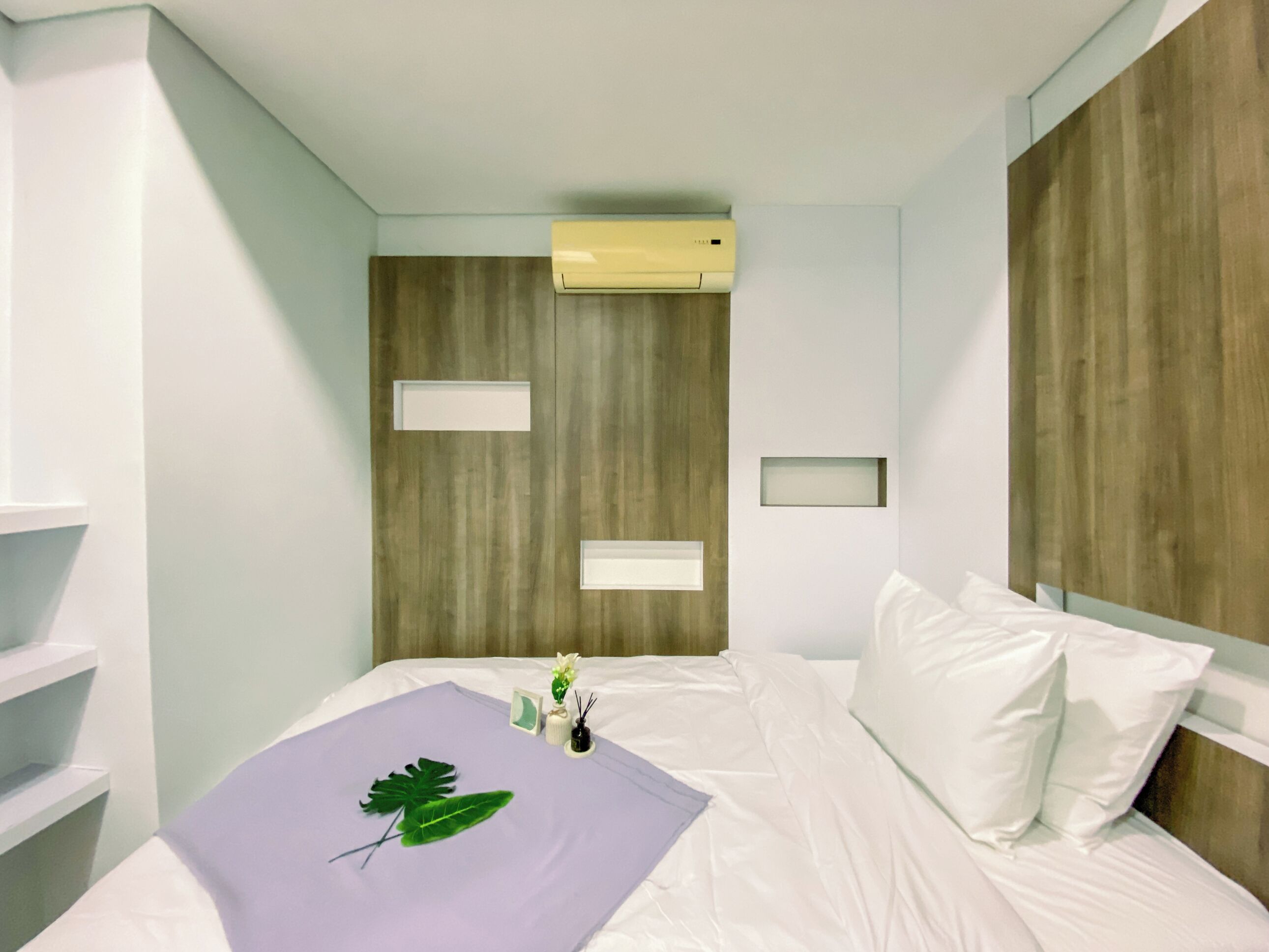 A modern hotel room with a bed, a wooden headboard, a wall-mounted air conditioner, and a small bedside table with a vase and a plant.