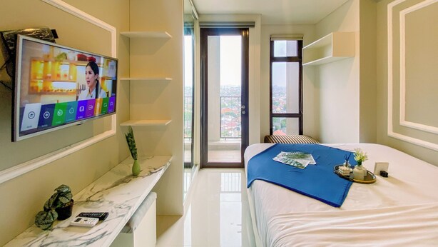 Ett modernt hotellrum med en stor TV på väggen, en säng med ett blått överkast, ett litet bord med en vas och en fjärrkontroll, och utsikt över staden genom fönstret.