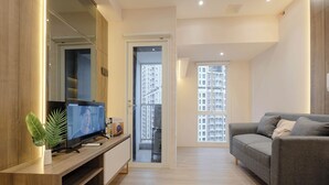 Un salon moderne doté d’un téléviseur à écran plat, d’un canapé gris, d’un plancher en bois et offrant une vue sur un immeuble de grande hauteur à travers une porte vitrée.