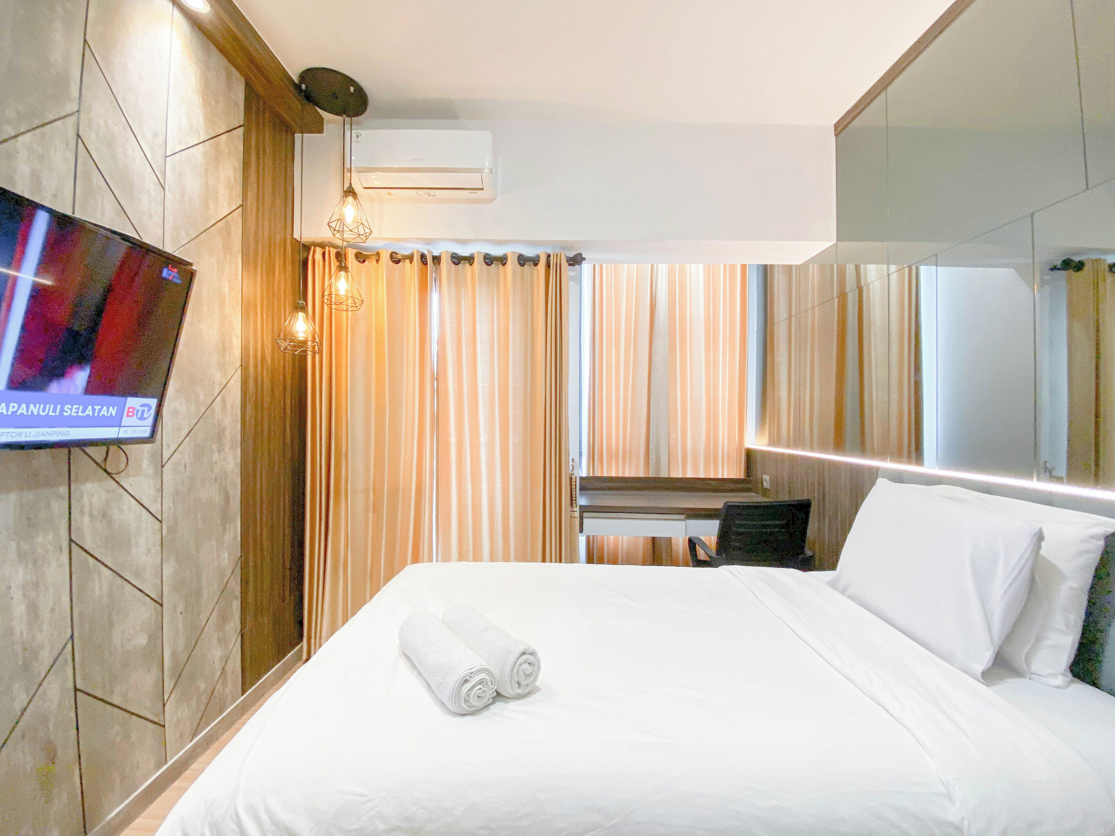 Quarto de hotel com cama, televisão na parede, escrivaninha e espelho.